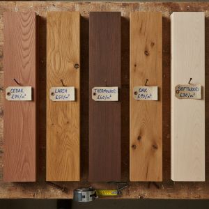 Five timber cladding samples — cedar, larch, thermowood, oak and softwood — standing on a workbench, each with a handwritten price-per-square-metre tag