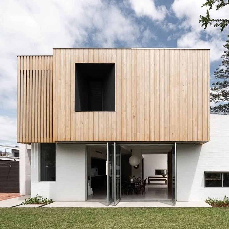 Modern two-story house with light wood cladding above white brick, large open glass doors revealing a dining area inside.
