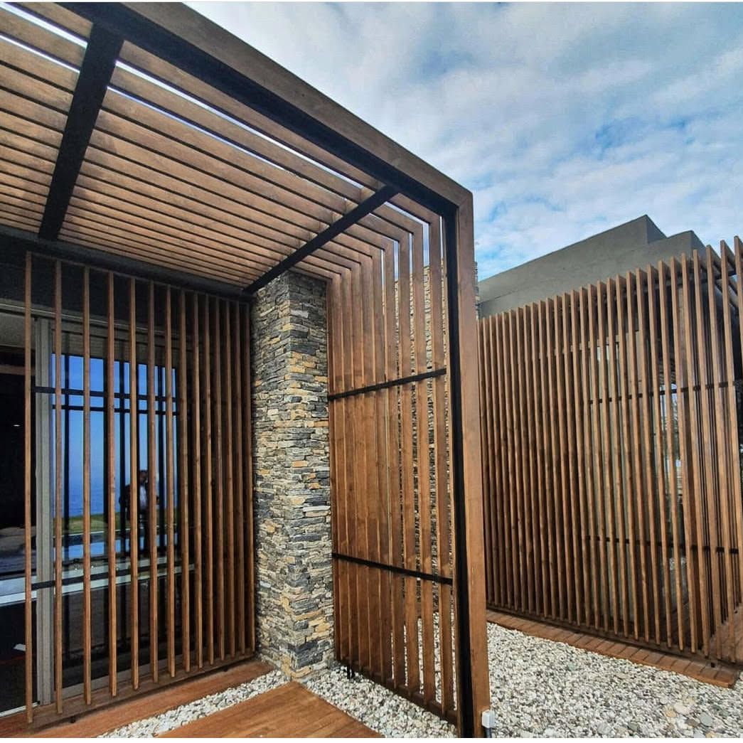 Modern exterior wooden slat fence with a stacked-stone pillar and gravel ground under a blue sky; outdoor architectural feature.