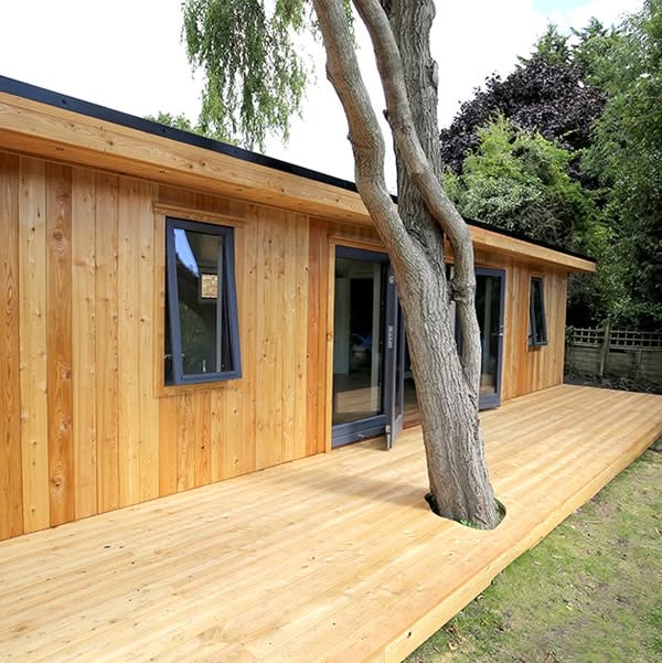 A wooden house with large glass doors and windows, built on a wooden deck that surrounds a tree growing through the deck edge.
