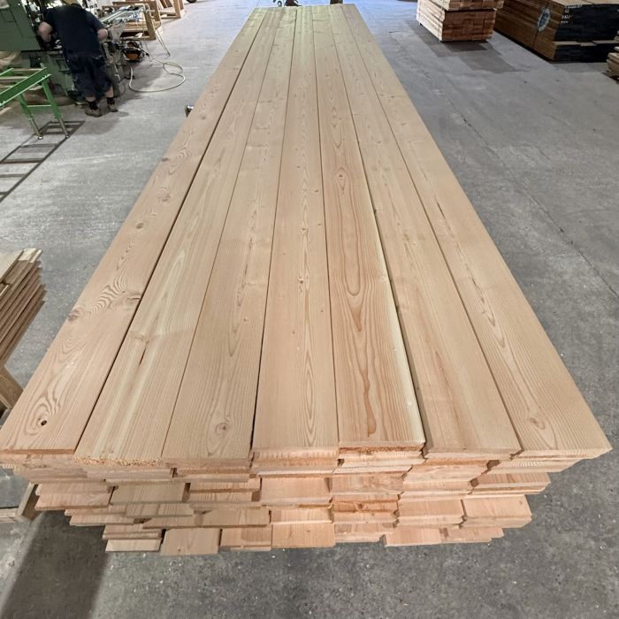Stack of long wooden planks neatly piled in a lumberyard, with a worker at a machine in the background to the left and other lumber in the distance.